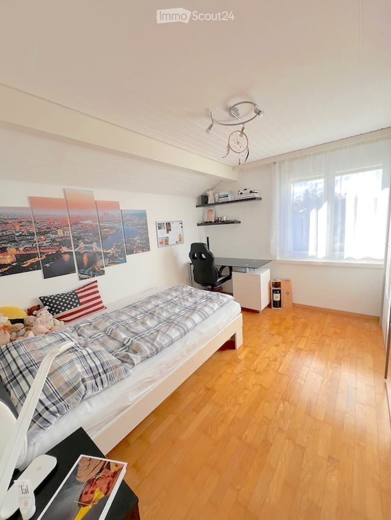 Cozy bedroom with wooden flooring, a single bed with plaid bedding, a desk with a chair, shelves with books and decor, and large windows with white curtains.
