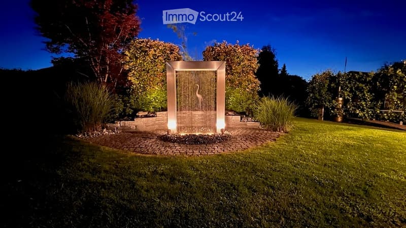 A modern outdoor water feature with a sleek, illuminated design, surrounded by well-maintained lawn and trees.