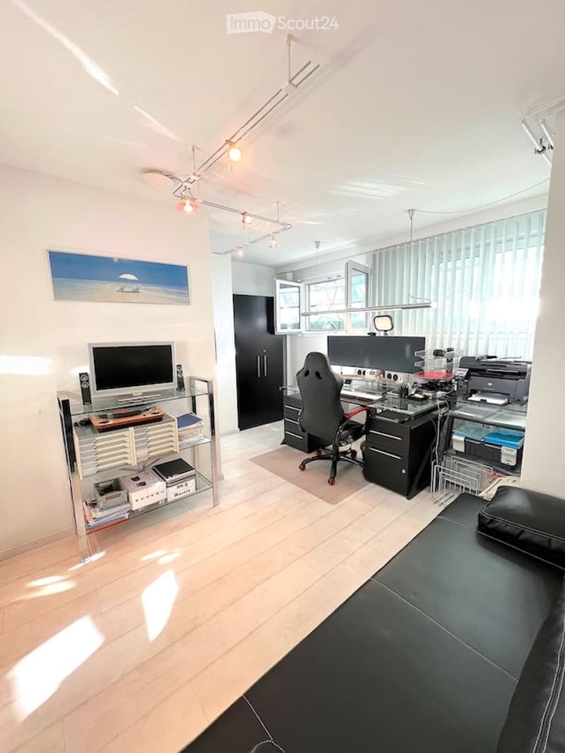 Modern office space with a large desk, computer setup, ergonomic chair, and a comfortable black leather sofa. The room features sleek white walls, large windows with blinds, and a stylish shelving unit with books and office supplies.