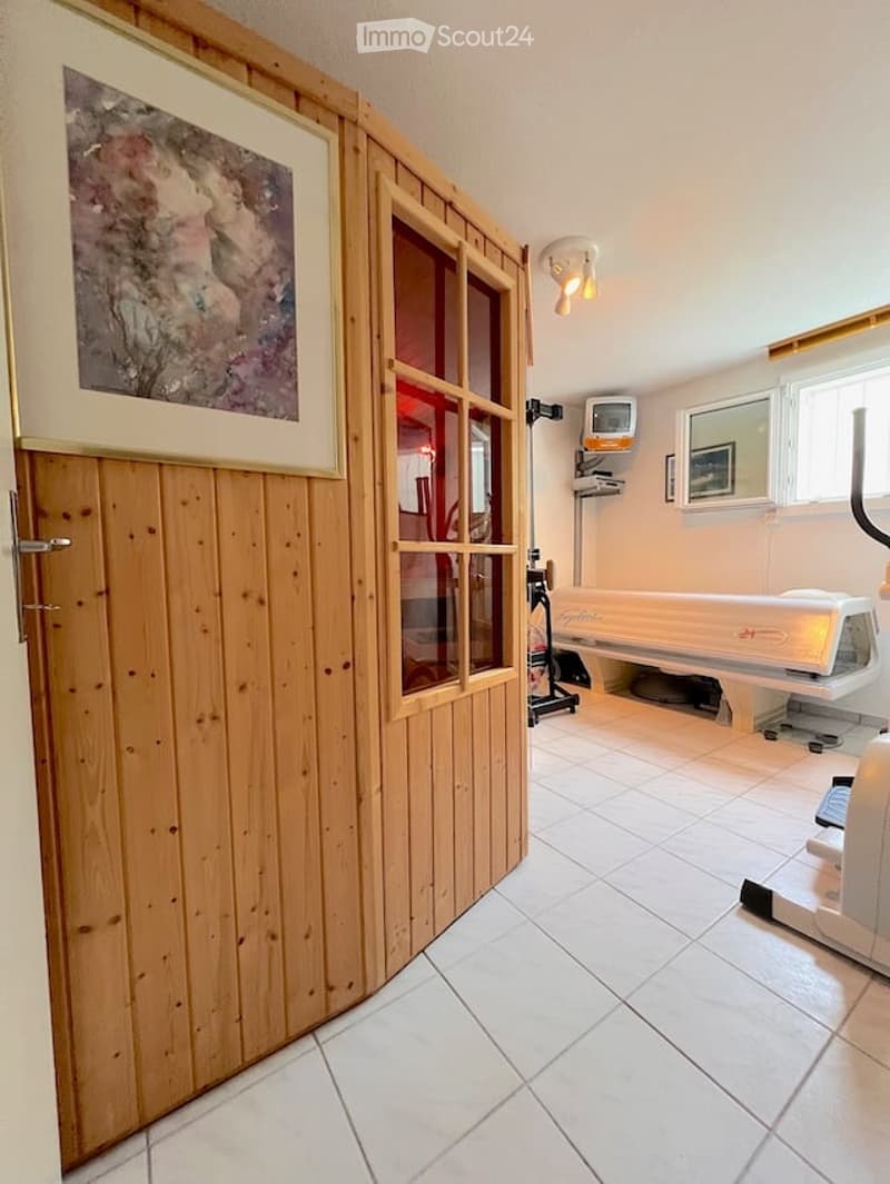A small, well-lit room with a tiled floor and wooden paneling. It features a tanning bed, a mirror, a small screen, and a picture frame on the wall.