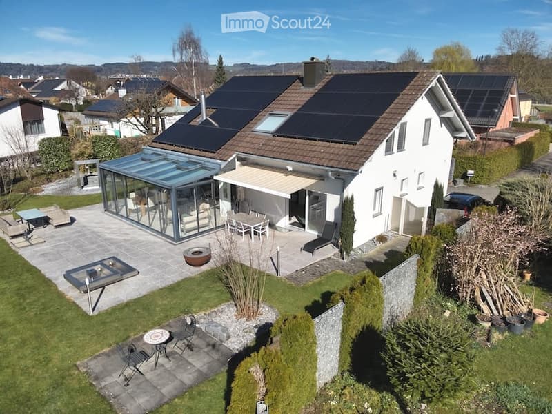 Modern two-story house with solar panels on the roof, surrounded by a well-maintained garden and outdoor seating areas.