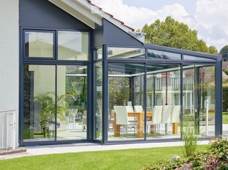 A modern glass-enclosed patio attached to a house, featuring a wooden dining table with white chairs, surrounded by greenery and lawn.