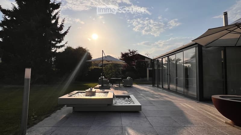 Modern outdoor patio with a water fountain, lounge chairs, umbrella, and sleek glass-walled building.