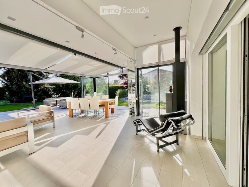 Bright, modern living and dining area with large glass walls, white furniture, and a sleek black leather chair. The space features a fireplace and a view of the garden.