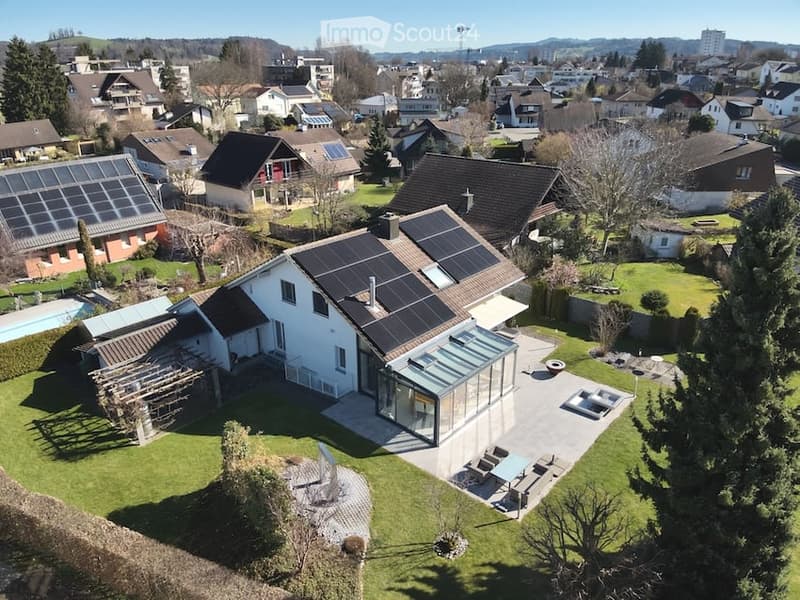 Aerial view of a modern two-story house with solar panels on the roof, surrounded by a well-maintained garden and a swimming pool in the backyard.