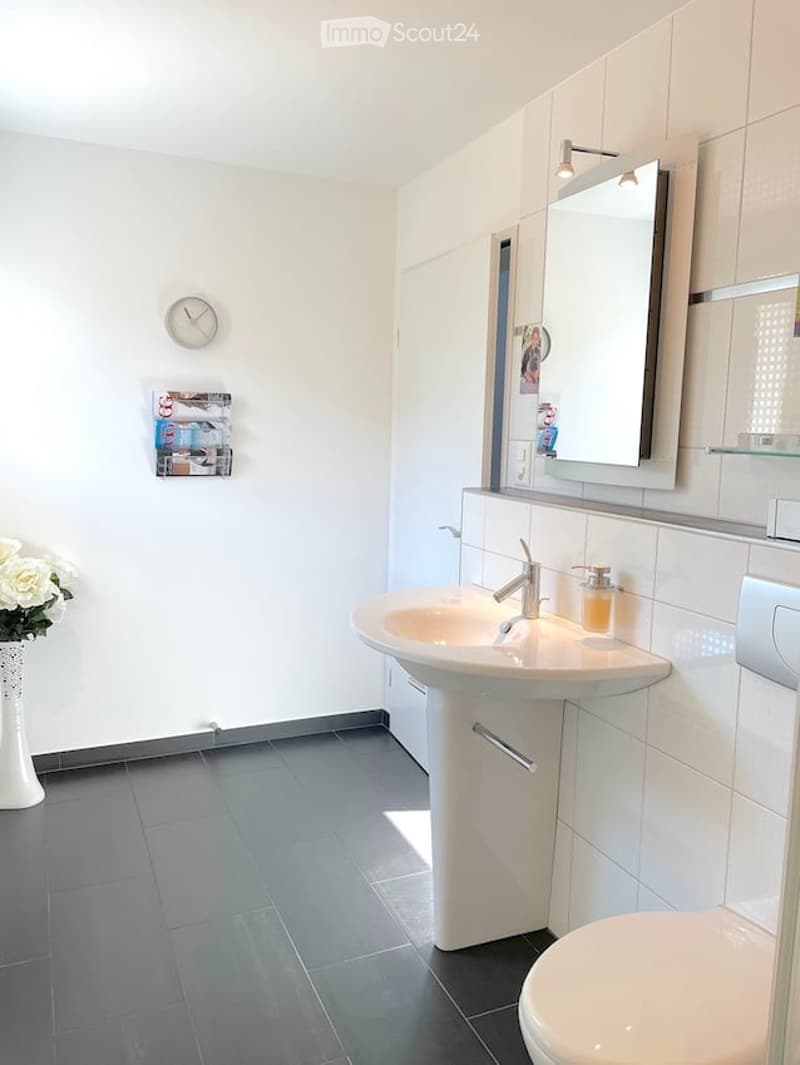 Modern bathroom with white tiles, a double sink, a large mirror, a toilet, a flower vase, and a clock on the wall.