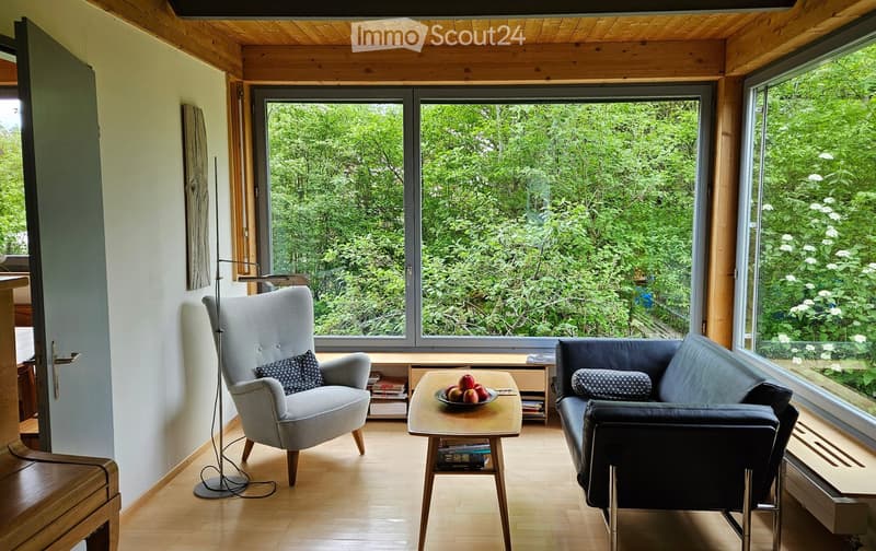Living Room. Bright living room with large windows overlooking a lush green garden, featuring a grey armchair, a dark blue sofa, a wooden coffee table with a bowl of fruit, and a floor lamp.