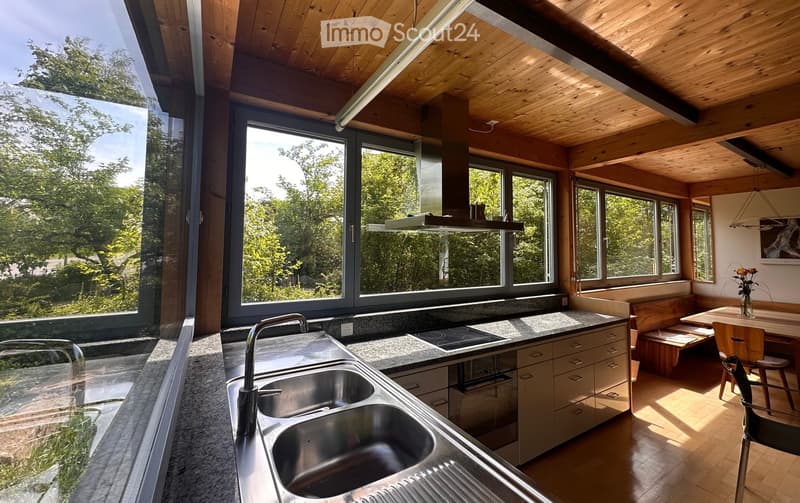Kitchen. Modern kitchen with stainless steel sink, granite countertops, built-in stove, and large windows overlooking a lush green landscape. The space features wooden cabinetry, a dining area with a bench and chairs, and a sleek, contemporary design.