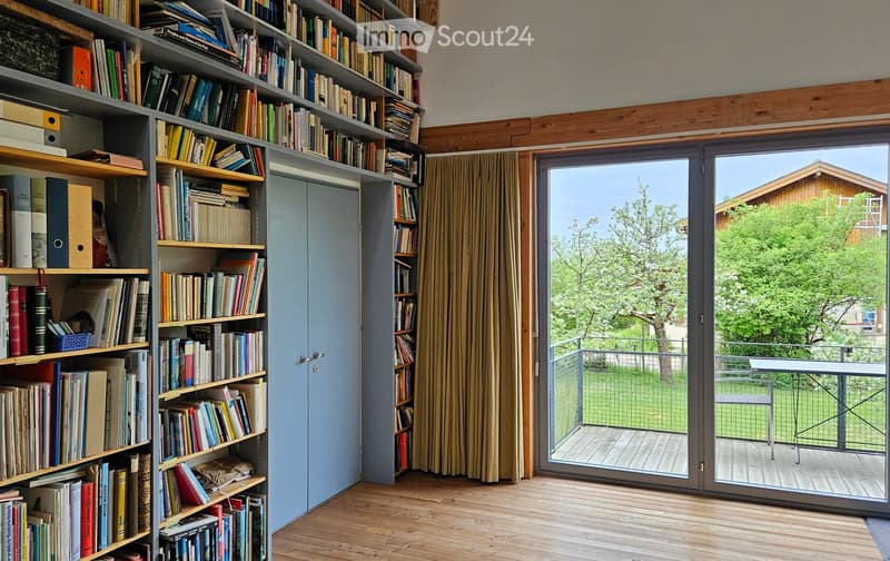 Furnished Room. A modern, well-lit room with wooden floors, large bookshelves filled with books, a sliding glass door leading to a balcony with outdoor furniture, and a view of greenery.