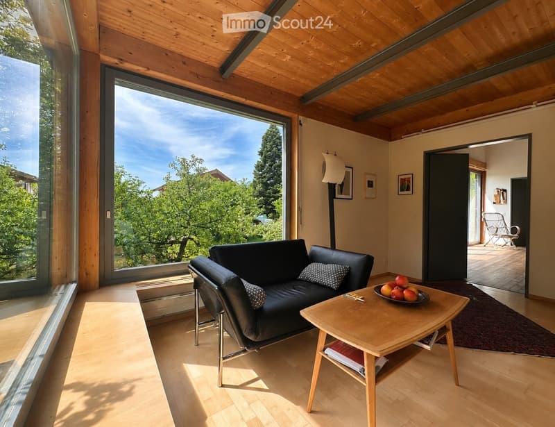 Living Room. Bright living room with wooden ceiling and floor, large window with view of greenery, black leather sofa, wooden coffee table with fruit bowl, and a modern floor lamp.