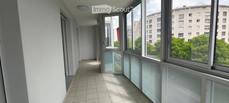 A modern, well-lit hallway with tiled flooring and large windows offering a view of the city and greenery outside.