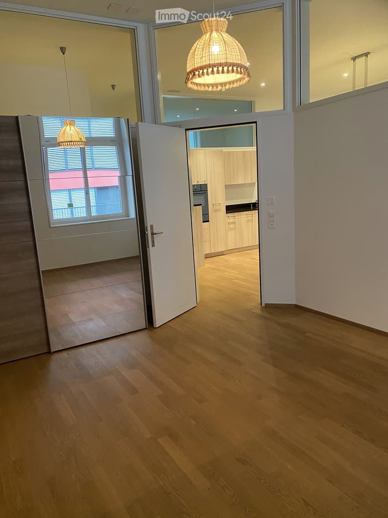 Empty room with wooden floor, white walls, large windows, open door, large mirror, kitchen visible through the door