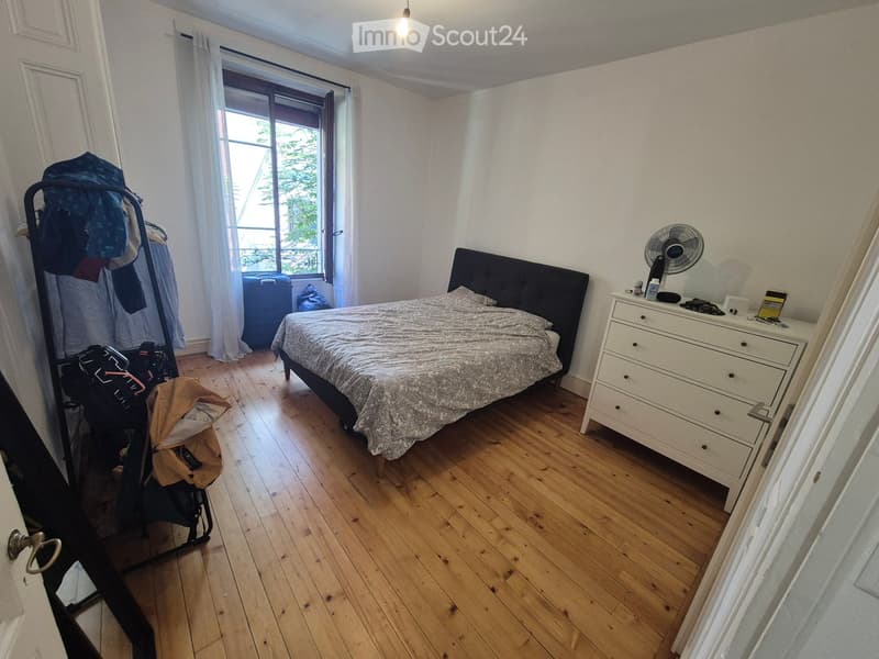 Bedroom with wooden flooring, a bed with a patterned duvet, a nightstand with a fan, a clothing rack with clothes, and a suitcase near the window.
