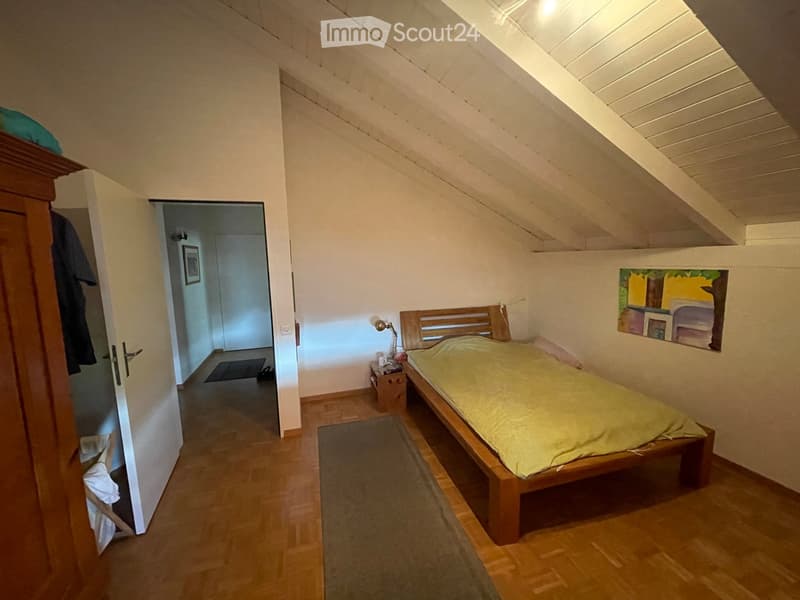 Cozy attic bedroom with wooden bed, nightstand, lamp, and painting on the wall. Slanted wooden ceiling and wooden flooring.