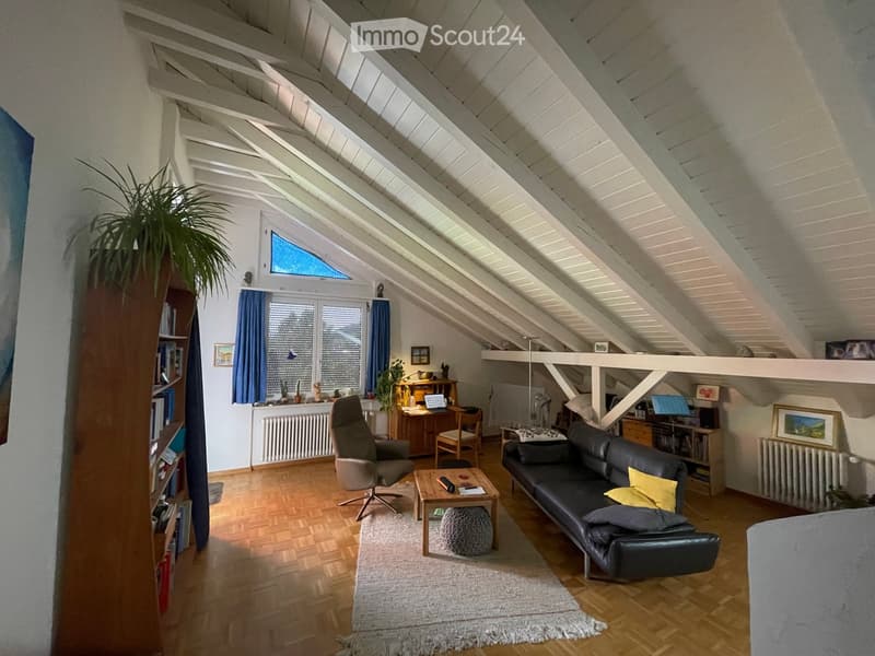 Bright living room with wooden flooring, slanted white ceiling beams, large windows, and skylight. Features a black leather sofa, wooden coffee table, armchair, desk with laptop, bookshelf, and decorative plants.