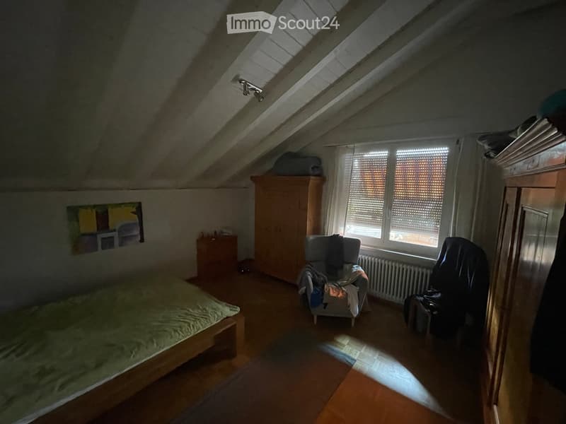 Cozy attic bedroom with wooden floors, slanted ceilings, and large windows. Features a bed, a chair with a blanket, a wardrobe, and a radiator.