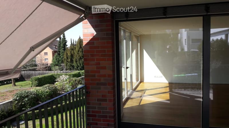 Balcony with wooden flooring, glass sliding doors, and a view of a garden with bushes and trees.