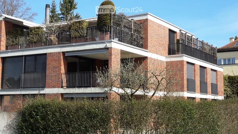 Modern brick apartment building with multiple balconies and greenery on the roof.