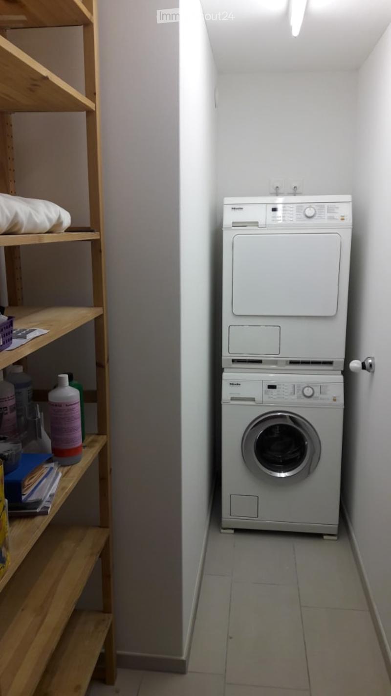 Compact bathroom with a stacked washer and dryer unit, wooden shelf with cleaning supplies and a pillow, white tiled floor, and bright lighting.