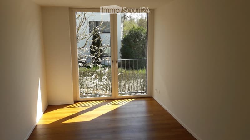 Empty room with wooden flooring and large glass sliding doors leading to a balcony area. Natural light streams in, highlighting the clean and minimalist design.