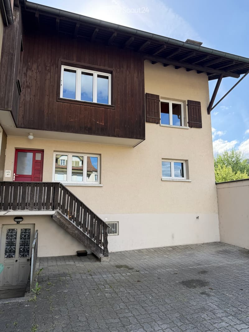 Two-story residential building with a wooden balcony, beige walls, and multiple windows. Features include a red front door, a staircase leading to the balcony, and a paved ground area.