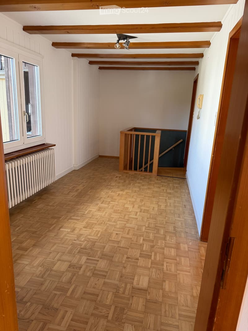 Empty room with wooden herringbone flooring, white walls, exposed wooden beams on the ceiling, and a radiator under a window. A wooden staircase railing is visible in the background.