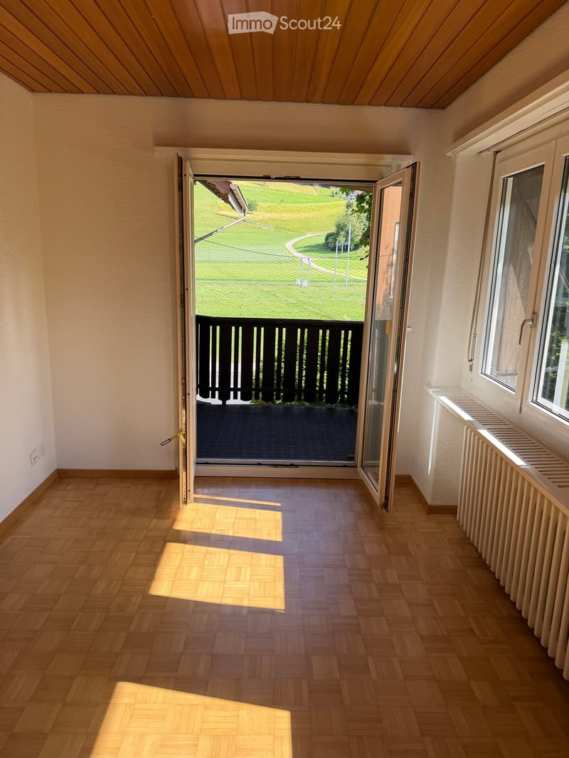 Empty room with wooden flooring, white walls, and a large open sliding door leading to a balcony with a view of a green lawn and trees.