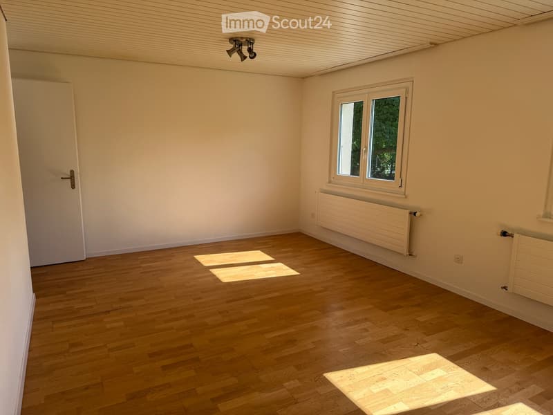 Empty room with wooden flooring, white walls, a single window, and a door. Natural light streams in, casting shadows on the floor.