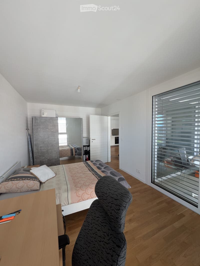 Modern bedroom with a large bed, patterned bedding, a wooden desk, a comfortable chair, and a large mirror. The room features wooden flooring and large windows with blinds.