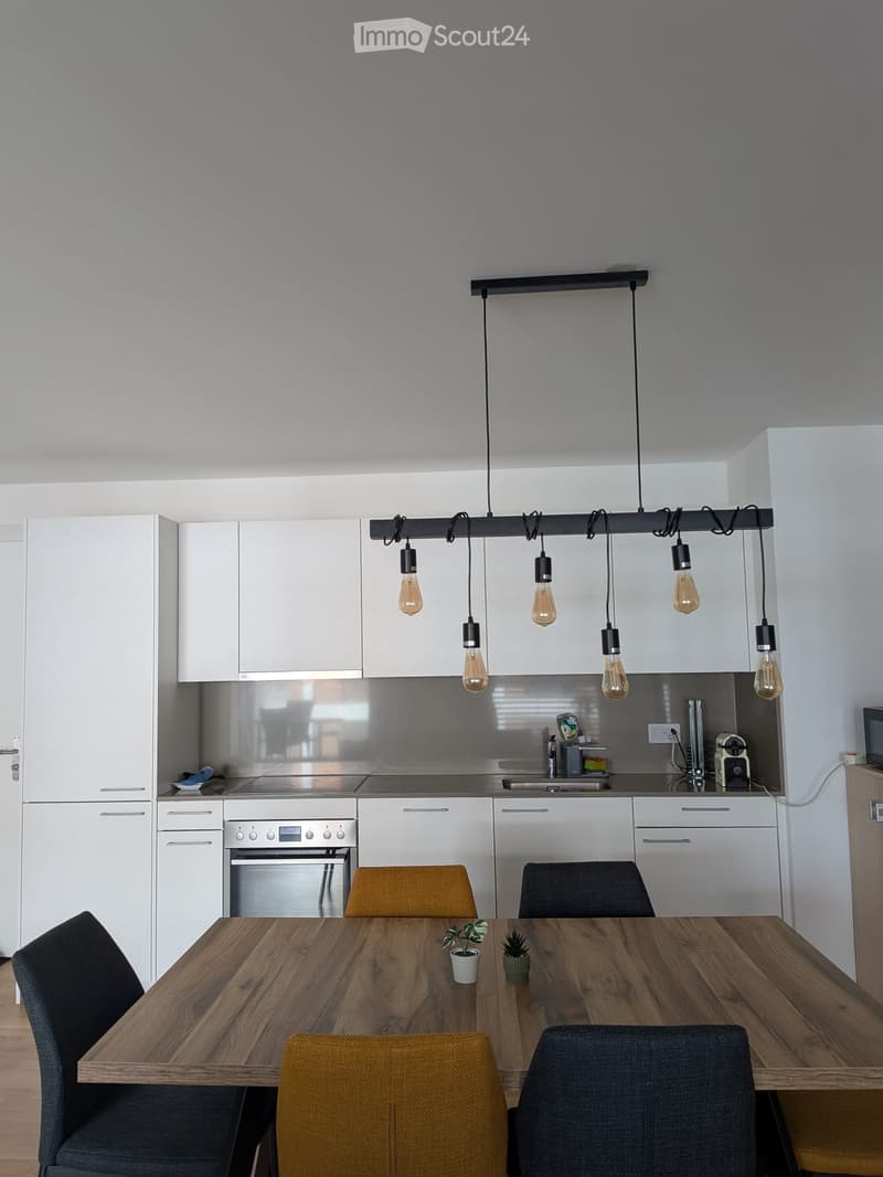 Modern kitchen with white cabinets, stainless steel appliances, a wooden dining table with four chairs, and pendant lighting.