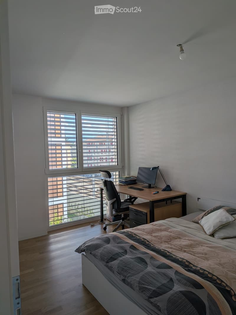 Modern bedroom with a double bed, desk, chair, computer, and lamp. Large windows with blinds offer natural light and a view of the city.