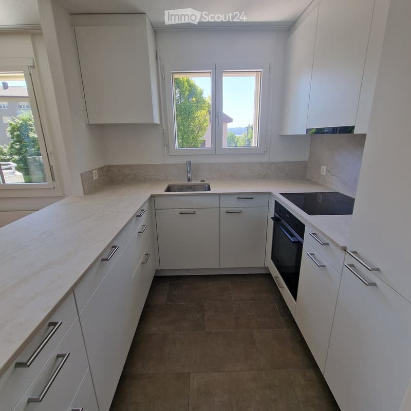 Modern kitchen with white cabinets, marble countertops, and stainless steel appliances including a sink and oven. Large windows provide natural light.