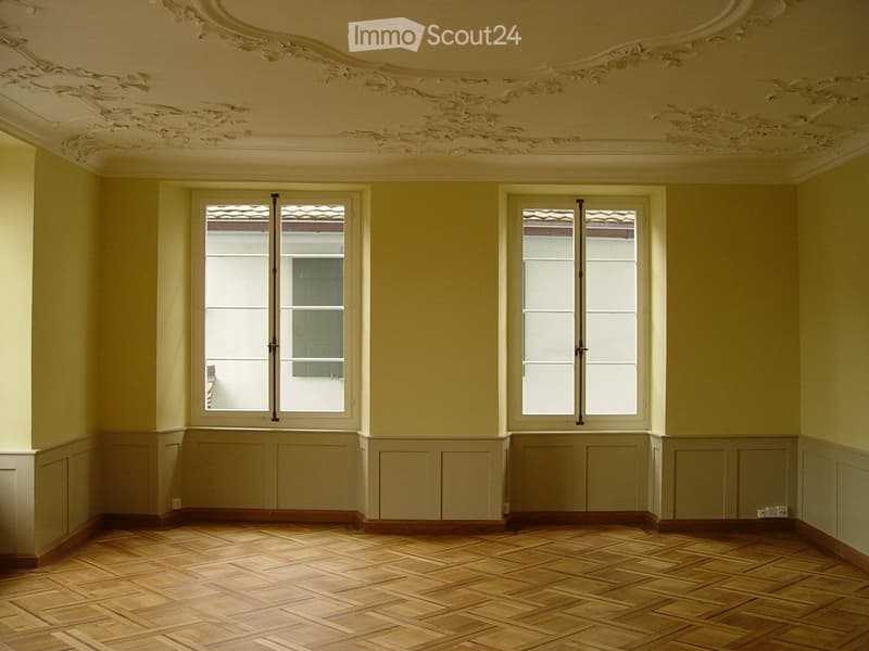 Empty room with wooden parquet flooring, light yellow walls, and two large windows with shutters. The ceiling features ornate plasterwork.