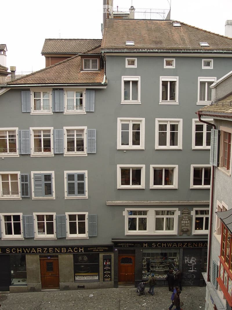 Four-story building with grey facade, multiple windows with shutters, and commercial storefronts on the ground floor.