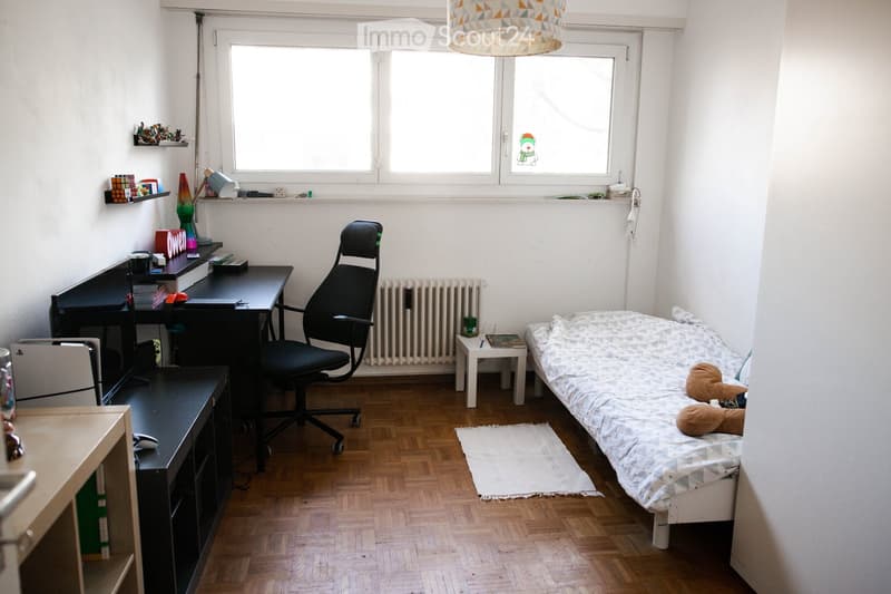 Cozy bedroom with a single bed, desk, chair, and shelves. Features wooden flooring, large windows, and a small rug.