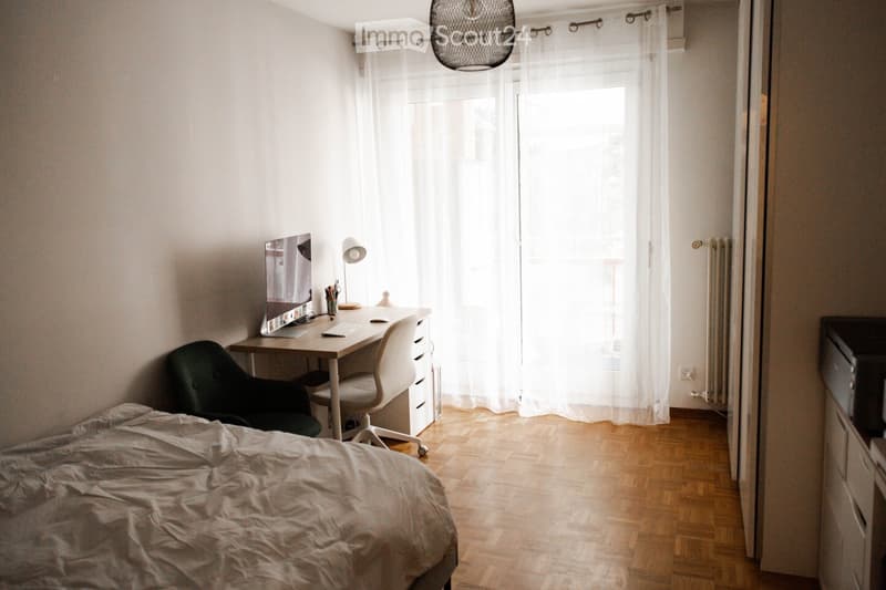 Cozy bedroom with a bed, desk, and chair. The room features wooden flooring, white walls, and large windows with sheer curtains.