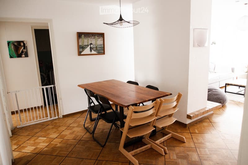 Dining area with a wooden table and chairs, pendant light, artwork on walls, and an adjacent living space with a sofa and ottoman.