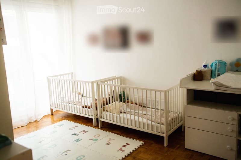 Brightly lit nursery with a white crib, changing table, and colorful toys. Wooden flooring and a white rug with letters.