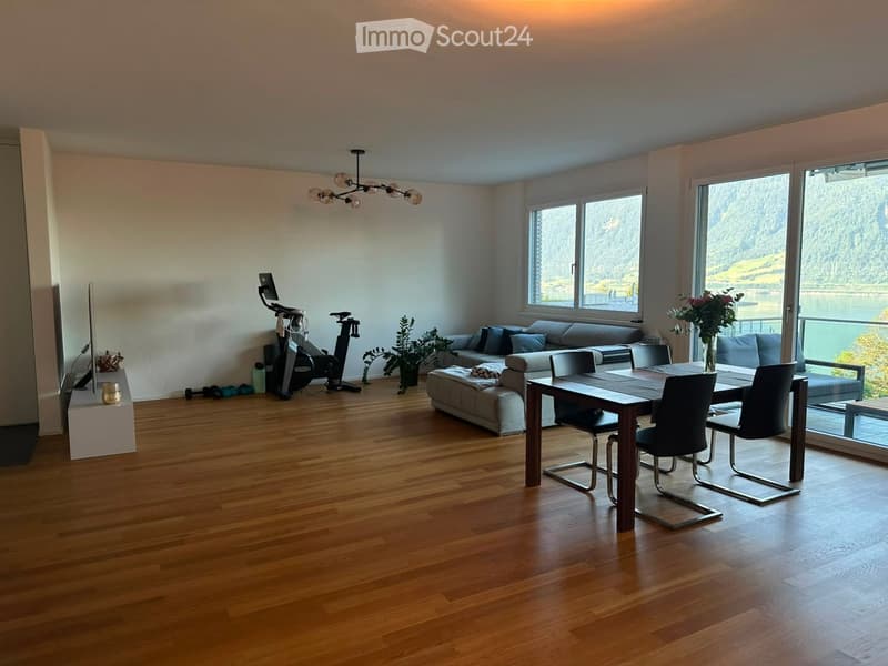 Open-plan living and dining area with wooden flooring, large windows offering a scenic view, a sofa with an ottoman, a dining table with chairs, exercise bike, screen, and decorative plants.