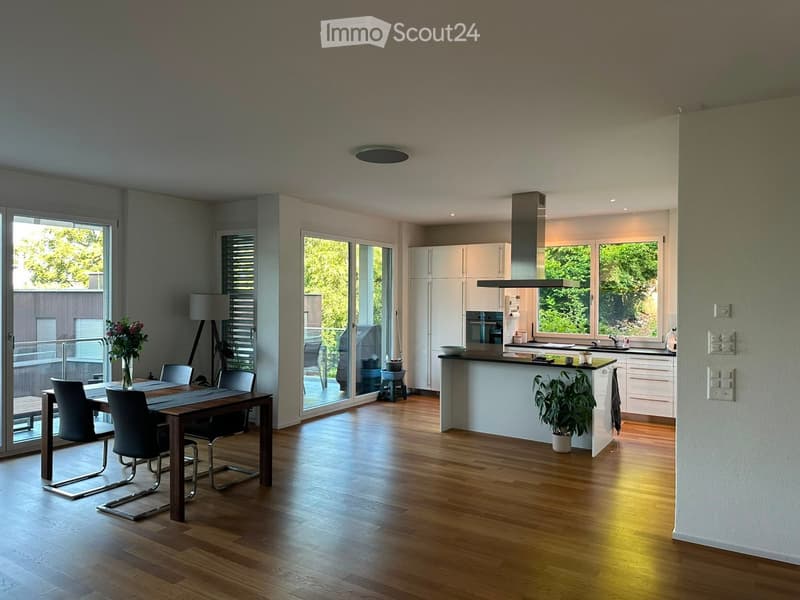Open-plan kitchen and dining area with wooden flooring, white cabinets, a central island, and large windows overlooking greenery.