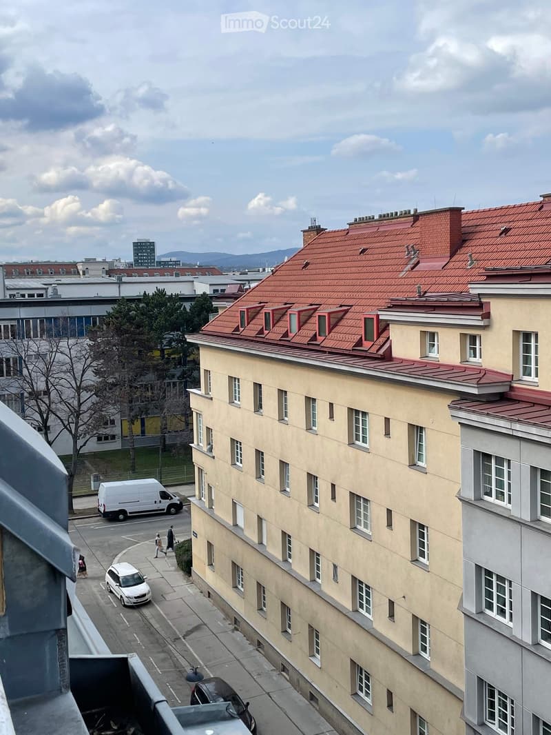Multi-story residential building with red-tiled roof and multiple windows, viewed from a balcony overlooking a street with parked cars and pedestrians.