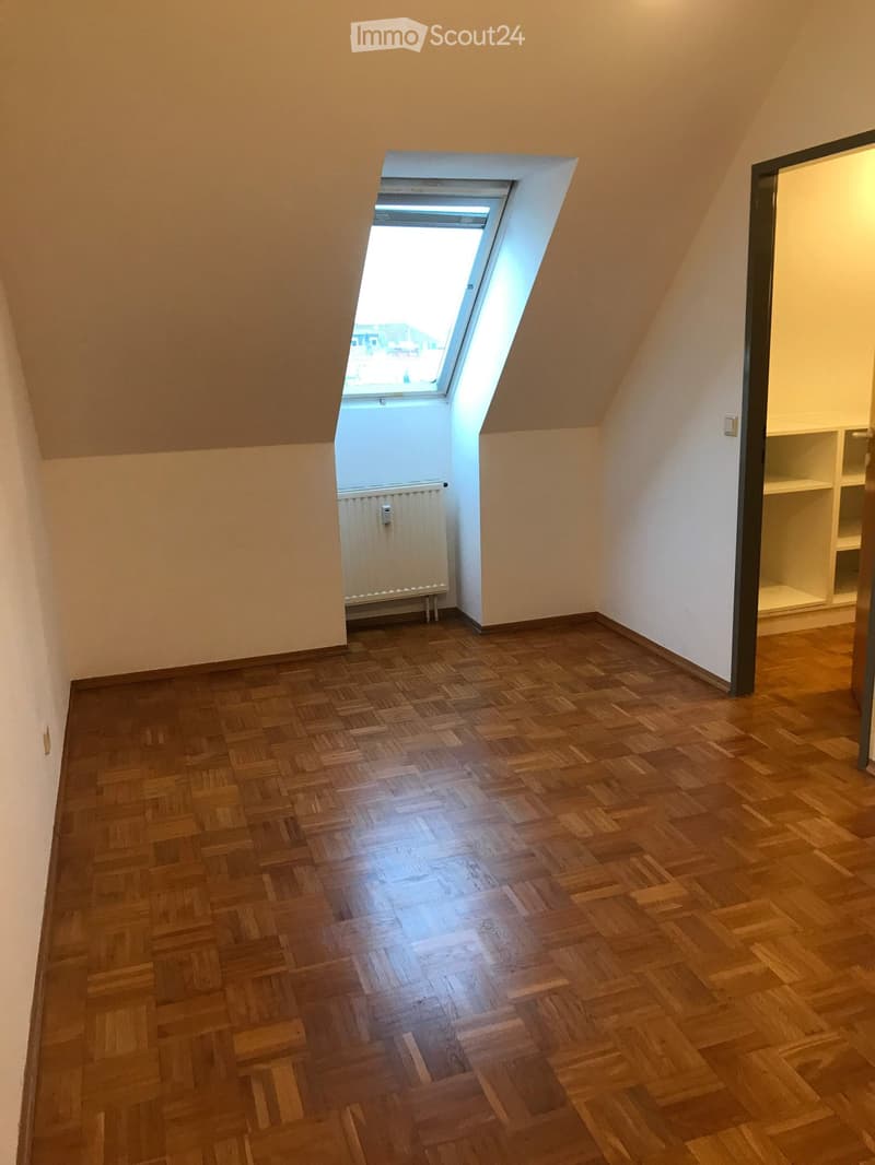 Empty attic room with wooden parquet flooring, white walls, and a skylight window. A radiator is visible near the window, and built-in shelving is on the right side.