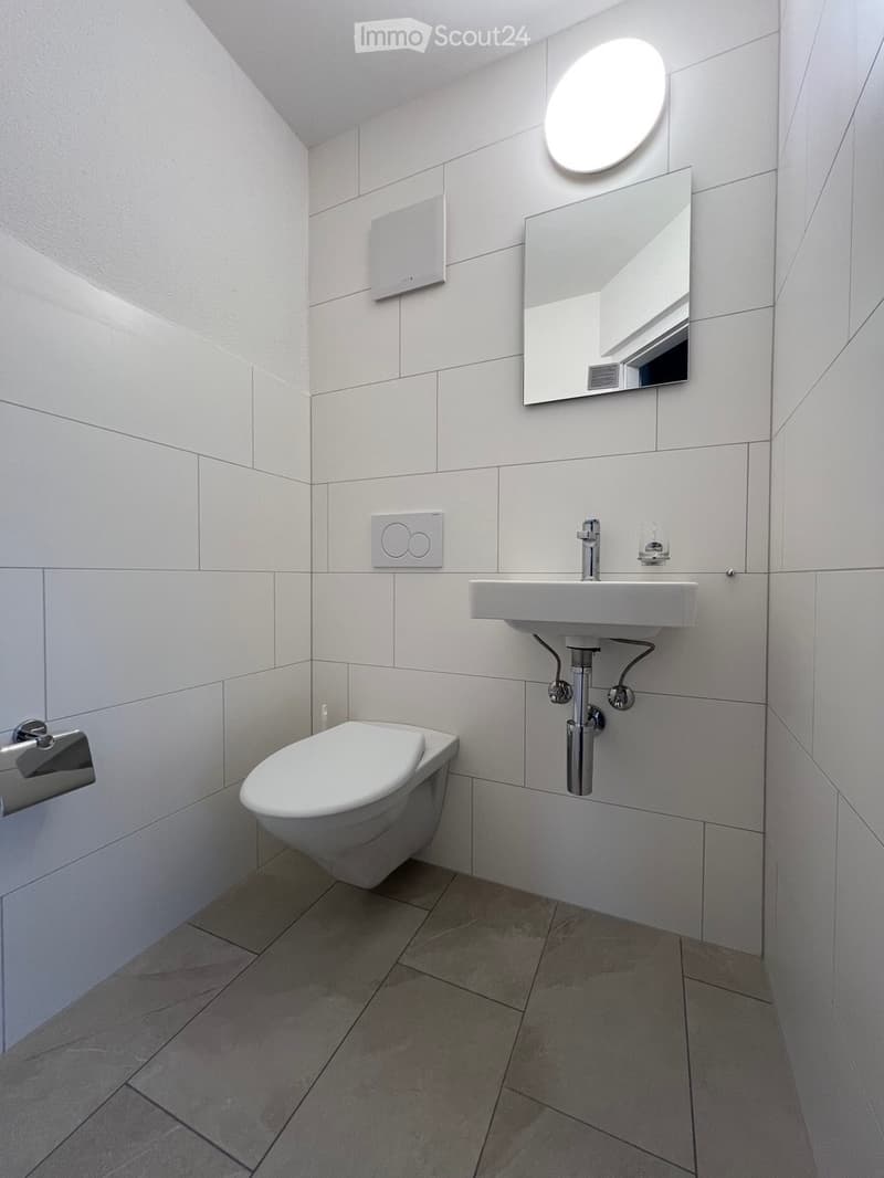 Modern bathroom with white tiled walls and floor, featuring a toilet, sink, mirror, and round light fixture above the mirror.