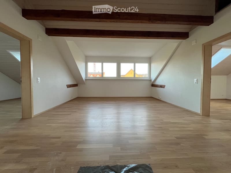 Empty attic room with wooden floors, exposed wooden beams, and large windows letting in natural light.