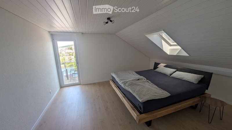 Modern bedroom with wooden floor, large window offering a scenic view, bed with dark bedding, nightstand, and small stool.