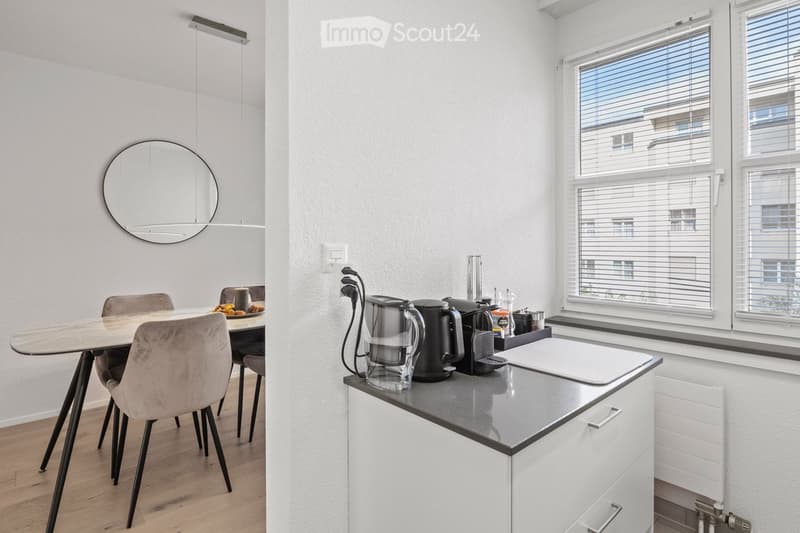 Modern kitchen and dining area with white cabinets, grey countertop, dining table with chairs, and various appliances.