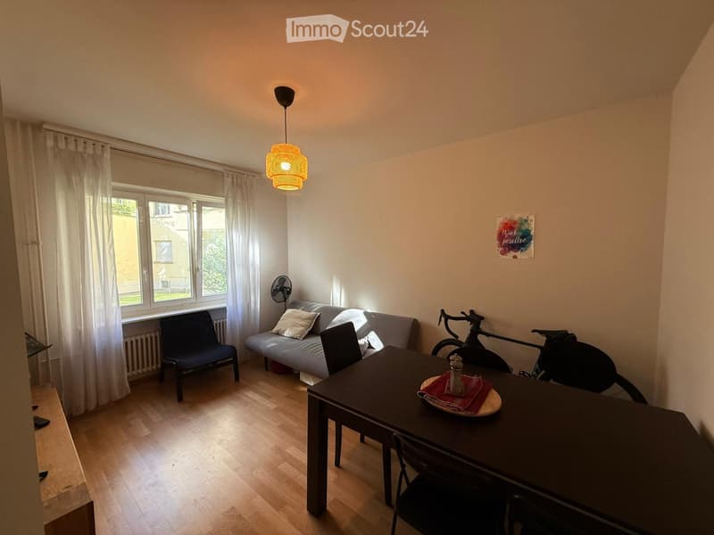 A modern, well-lit living space with a sofa, dining table, chairs, a fan, and a bicycle. The room features wooden flooring, large windows with white curtains, and a decorative painting on the wall.