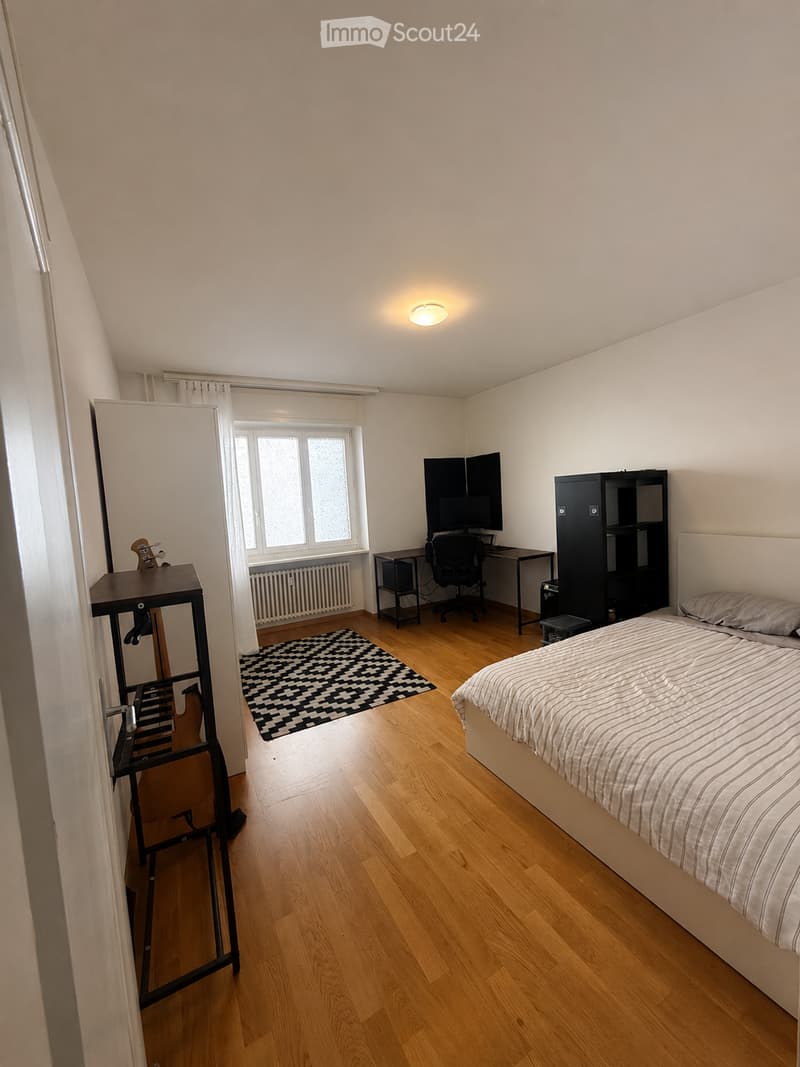 Bright bedroom with wooden flooring, large window with curtains, bed, desk with chair and screen, cabinet, mirror, and small table with items.
