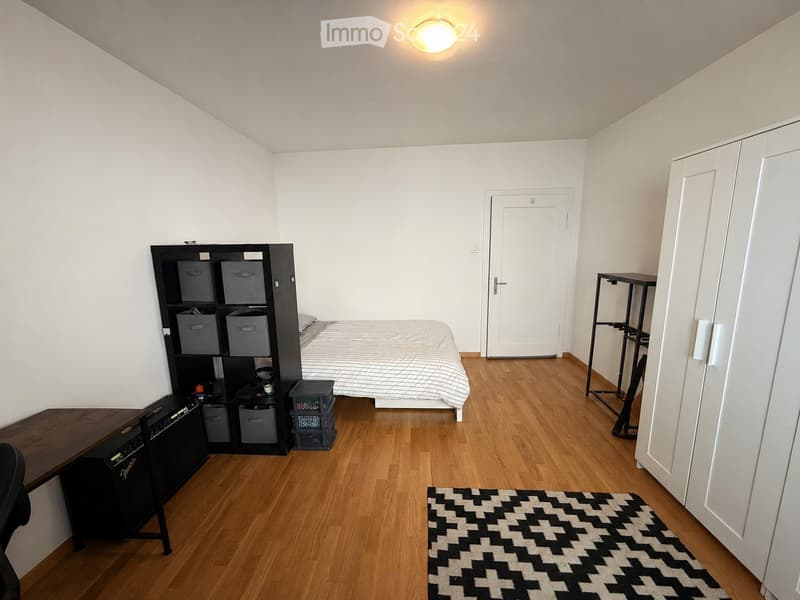 A small, furnished bedroom with a bed, desk, chair, storage unit, and a rug on a wooden floor.