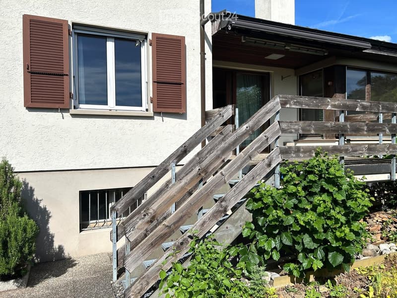 Two-story house with beige exterior, large windows with brown shutters, wooden staircase leading to a balcony, and greenery in the foreground.